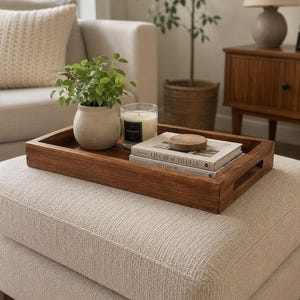 May include: A rectangular dark brown wooden serving tray with cutout handles. Inside the tray sits a small off-white ceramic pot with a green plant, a clear glass candle with a black label, and two stacked books titled Live Beautifully.