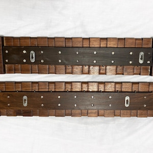 May include: Two wooden wall shelves with a dark brown finish. Each shelf has a central dark brown panel with metal keyhole hangers and is framed by a series of small, square wooden blocks. The shelves are designed for wall mounting.