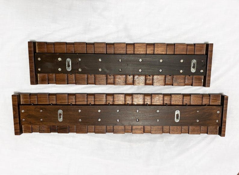 May include: Two wooden wall shelves with a dark brown finish. Each shelf has a central dark brown panel with metal keyhole hangers and is framed by a series of small, square wooden blocks. The shelves are designed for wall mounting.