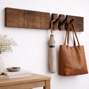 May include: A wooden wall-mounted coat rack with three flip-down hooks. A tan umbrella and a brown leather tote bag hang from the hooks. A vase of white flowers sits on a wooden table next to the rack.