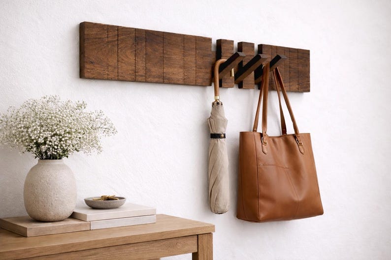 May include: A wooden wall-mounted coat rack with three flip-down hooks. A tan umbrella and a brown leather tote bag hang from the hooks. A vase of white flowers sits on a wooden table next to the rack.