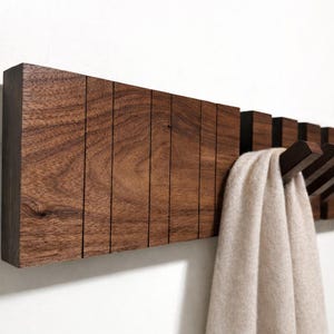 Puede incluir: Un perchero de madera marrón oscuro con múltiples ganchos. El perchero está montado en una pared blanca y sostiene una bufanda beige. La veta de la madera es visible y el diseño es moderno y minimalista.