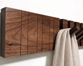 May include: A dark brown wooden wall-mounted coat rack with multiple hooks. The rack has a rectangular design with vertical grooves and a scarf hanging from one of the hooks. The scarf is a light beige colour.