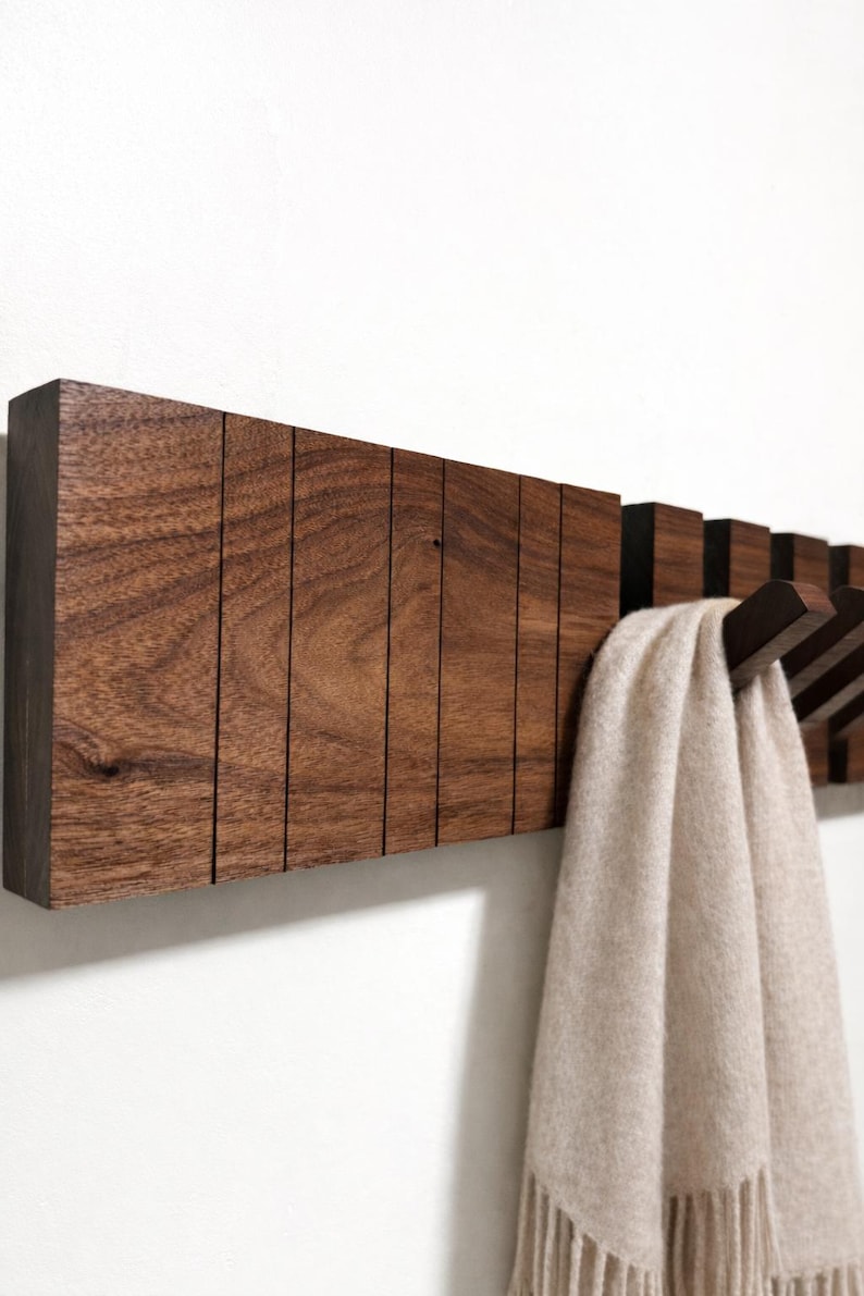 May include: A dark brown wooden wall-mounted coat rack with multiple hooks. The rack has a rectangular design with vertical grooves and a scarf hanging from one of the hooks. The scarf is a light beige color.