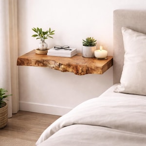 May include: A floating wall shelf made of natural light brown wood with a live edge, featuring a rough, organic front texture. The shelf holds a small potted green plant, a stack of two white books with black glasses on top, and a white candle.
