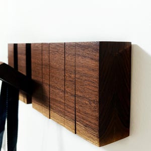 May include: A modern wooden wall-mounted coat rack with multiple hooks. The rack is made of dark brown wood and has a rectangular shape. A black belt hangs from one of the hooks. The rack is mounted on a white wall.
