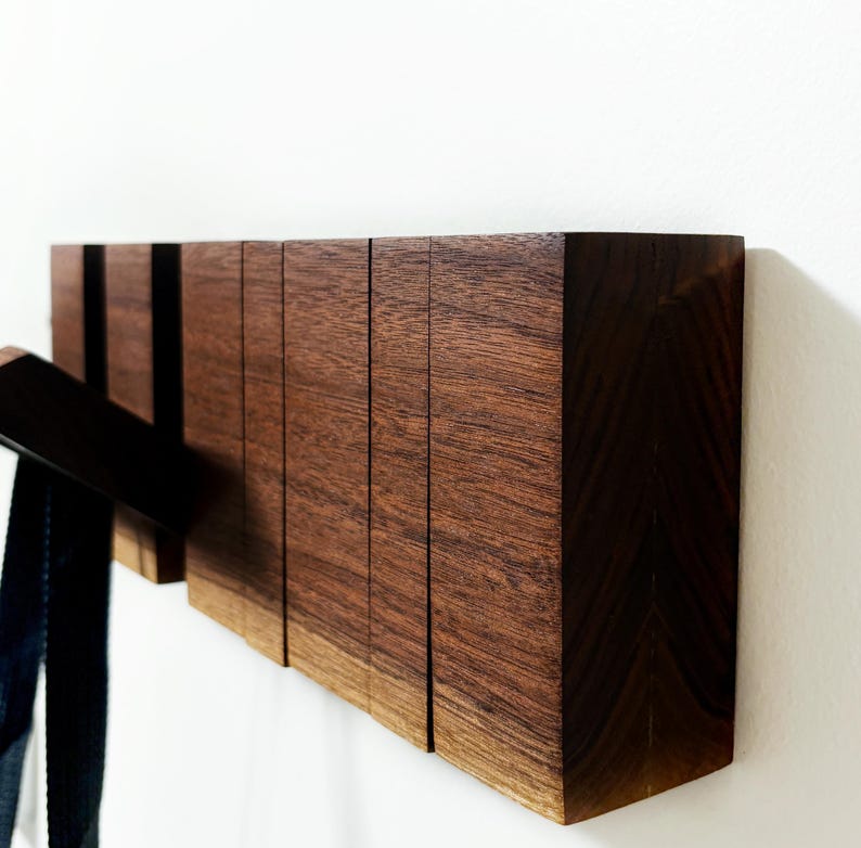 May include: A modern wooden wall-mounted coat rack with multiple hooks. The rack is made of dark brown wood and has a rectangular shape. A black belt hangs from one of the hooks. The rack is mounted on a white wall.
