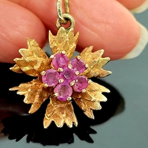 以下が含まれることがあります： ピンクの宝石がついた金のフラワーペンダントネックレス。ペンダントはテクスチャ加工された金で詳細に作られており、繊細なチェーンが付いています。