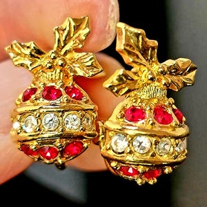 May include: A pair of gold-toned earrings with a festive design. Each earring features a rounded shape adorned with red and clear gemstones, topped with a decorative gold leaf element. The earrings are designed to resemble ornaments.
