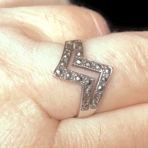 May include: A silver-toned ring with a zigzag design, set with small, dark gemstones. The ring is worn on a finger, showcasing its geometric shape and the sparkle of the stones. The ring's design is a bold, modern statement.