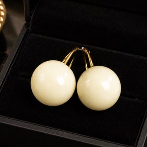 May include: A pair of cream-colored spherical earrings with gold-toned metal lever-back closures, presented inside a black velvet-lined jewelry box. The smooth, round beads have a glossy finish and are positioned together in the center of the box.