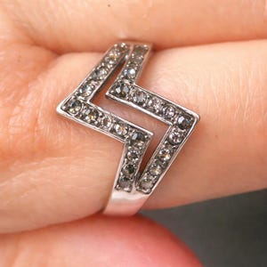 以下が含まれることがあります： 幾何学的なジグザグ模様のシルバーリング。小さな丸い薄茶色の宝石がちりばめられています。指にはめられており、ジュエリーの繊細なディテールが際立っています。