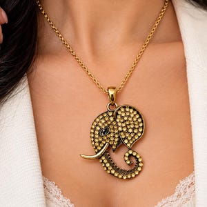 Op de afbeelding: Een goudkleurige ketting met een hanger in de vorm van een olifantenkop. De olifantshanger is ingelegd met talloze kleine, fonkelende gele kristallen en heeft een donkere steen als oog, hangend aan een eenvoudige gouden ketting.