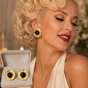 May include: Gold-toned earrings with a black center stone, presented in a gray velvet box. The earrings have a textured, circular design. The box is labeled "Timeless Beauty".