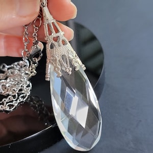 May include: A clear, faceted crystal pendant necklace with a silver-toned filigree cap and chain. The teardrop-shaped crystal reflects light, creating a sparkling effect. The necklace is displayed against a dark background.