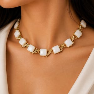Peut inclure: Un collier doré orné de pierres carrées blanches. Les pierres sont reliées par un motif délicat en forme de feuilles. Le collier est porté sur une chemise blanche.