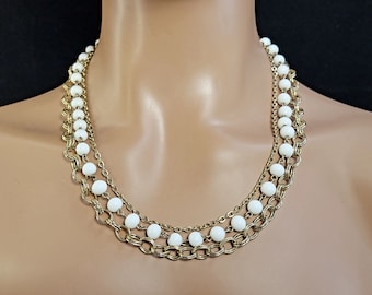 May include: A vintage necklace with three strands. The necklace features white, round beads and gold-tone chains. The chains have a mix of delicate and chunky links. The necklace is displayed on a mannequin.