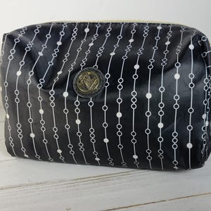 Op de afbeelding: Zwarte cosmetische tas met een gouden rits en een herhalend wit stippellijnenpatroon. Een gouden embleem is gecentreerd op de tas. De tas is rechthoekig met afgeronde hoeken.