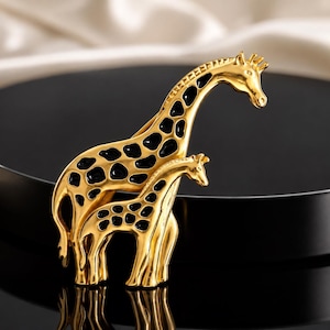 May include: A gold-toned metal brooch featuring two giraffes, one larger than the other, with black enamel spots. The giraffes are depicted standing side-by-side, with the taller giraffe positioned slightly behind the smaller one, both facing right.