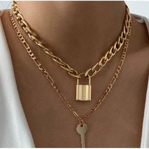 May include: Gold-toned layered necklace set. The top layer features a thick chain with a padlock pendant. The second layer is a thinner chain with a key pendant. The necklace is a stylish accessory.