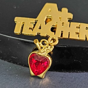以下が含まれることがあります： 「A+ TEACHER」の文字と、赤いハート型の宝石が付いたリンゴのチャームが付いた、金色の金属製ピン。このピンは、先生への感謝の贈り物やアクセサリーとして使われる可能性があります。
