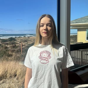 May include: White t-shirt featuring a red graphic of a cartoon character with a floral crown and dress. The short-sleeved shirt is worn by a person standing in front of a window with a view of the ocean and blue sky.