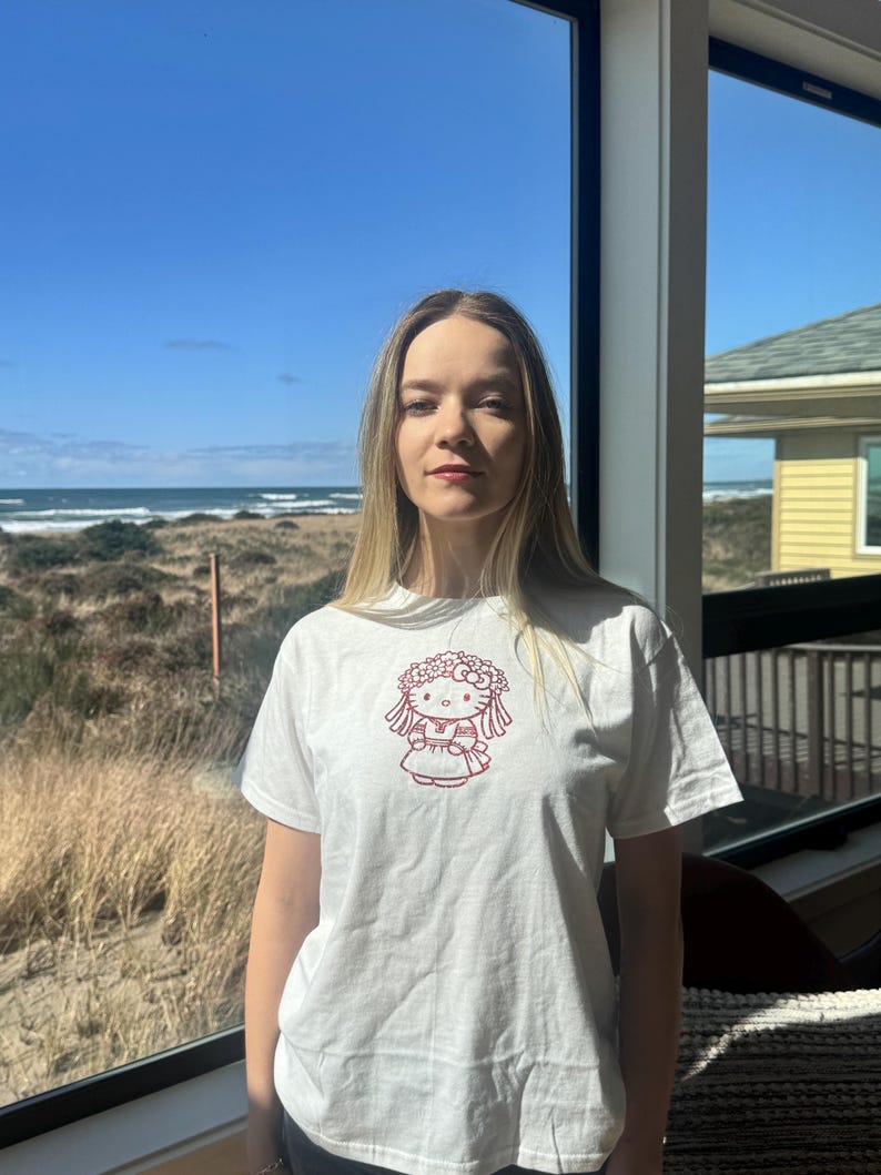May include: White t-shirt featuring a red graphic of a cartoon character with a floral crown and dress. The short-sleeved shirt is worn by a person standing in front of a window with a view of the ocean and blue sky.