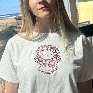 May include: White t-shirt featuring a maroon graphic of a cartoon character wearing a floral crown. The character is depicted in a dress and has long hair. The shirt is made of a soft material.