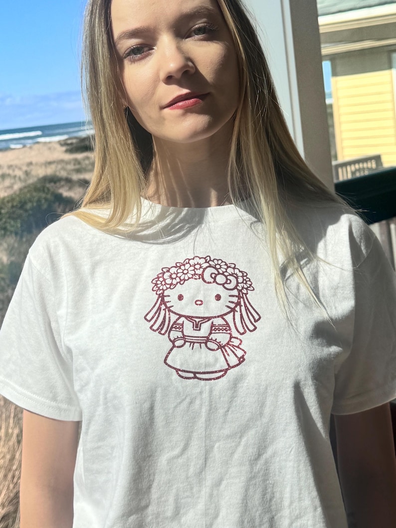 May include: White t-shirt featuring a maroon graphic of a cartoon character wearing a floral crown. The character is depicted in a dress and has long hair. The shirt is made of a soft material.