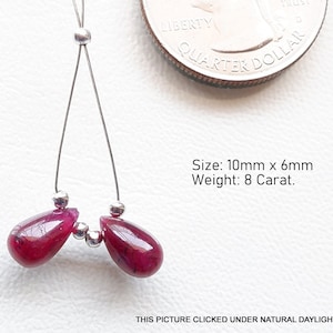 May include: Two teardrop-shaped ruby gemstones, each measuring 10mm x 6mm, strung on a silver wire with silver beads. The gemstones are a deep red colour and have a slight sparkle. The image includes the text "Size: 10mm x 6mm Weight: 8 Carat."