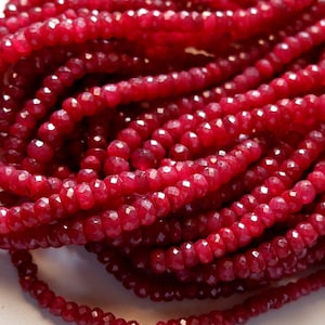 Op de afbeelding: Een streng dieprode robijn kralen, gefacetteerd en gepolijst, met een gouden kwast aan het einde. De kralen zijn geregen op een doorzichtige draad en zijn ongeveer 4 mm in diameter.