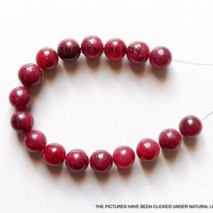 Op de afbeelding: Een streng van 18 ronde, rode robijn kralen, elk ongeveer 6 mm in diameter. De kralen zijn geregen op een dun, doorzichtig draad.