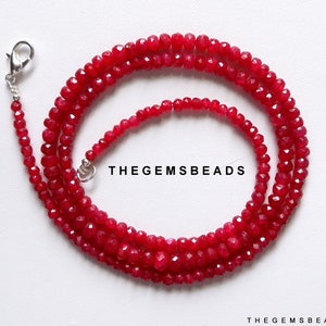 Op de afbeelding: Een streng rode edelsteen kralen met een zilveren sluiting. De kralen zijn facetgeslepen en hebben een glanzende afwerking. TheGEMSBeads is gedrukt op een witte achtergrond achter de kralen.