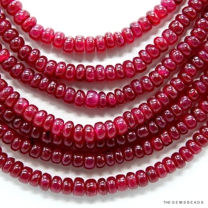 May include: A strand of deep red ruby beads, each bead is round and slightly elongated, creating a textured look. The beads are strung together to form a long necklace.