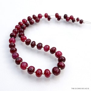 Puede incluir: Una hebra de cuentas de rubí rojo oscuro enhebradas en un alambre plateado. Las cuentas son redondas y lisas, con una ligera variación de tamaño. Las cuentas están dispuestas en un patrón graduado, con las cuentas más grandes en la parte inferior de la hebra.