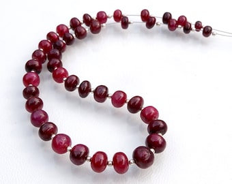 May include: A strand of deep red ruby beads strung on a silver wire. The beads are round and smooth, with a slight variation in size. The beads are arranged in a graduated pattern, with the larger beads at the bottom of the strand.