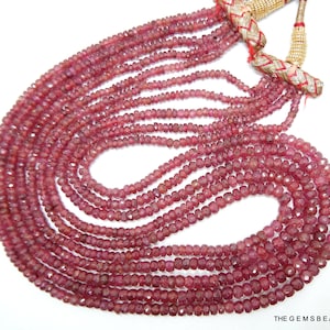 May include: A strand of pink ruby beads, faceted and round, strung on a thread. The beads are arranged in a circular pattern, with the ends of the strand meeting at the top of the circle. The beads are a deep pink color, and they have a slight sparkle to them.