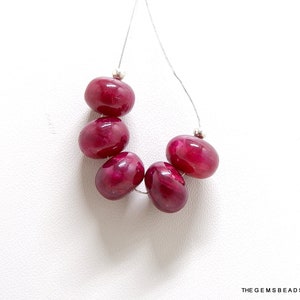 May include: Six deep red gemstone beads strung on a silver thread. The beads are oval shaped and have a smooth, polished finish.