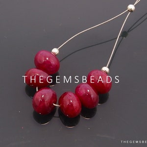 Może przedstawiać: Szereg czerwonych koralików z kamieni szlachetnych nawleczonych na srebrny drut. Koraliki mają owalny kształt i gładkie, polerowane wykończenie. W obrazie widoczny jest tekst "THEGEMSBeads".