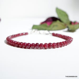 Pode incluir: Uma pulseira de contas vermelhas com fecho prateado. A pulseira é feita de pequenas contas redondas e polidas. O fundo é branco com uma flor e folhas desfocadas.