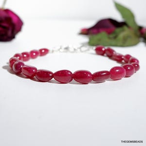 May include: A bracelet featuring polished, oval-shaped, deep red beads strung together with a silver clasp. The beads are evenly spaced, creating a smooth, continuous line. The background is a soft white, with a hint of a dark red rose.