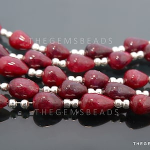 Puede incluir: Una hebra de cuentas de piedras preciosas de color rojo intenso, cada cuenta tiene forma de lágrima. Las cuentas están enhebradas en un alambre plateado con pequeñas cuentas espaciadoras plateadas entre cada cuenta de piedra preciosa. El texto "THEGEMSBeads" es visible en la imagen.