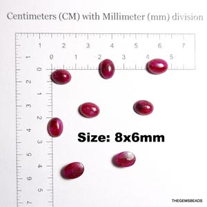 Peut inclure: Plusieurs pierres précieuses ovales, rouge foncé, d'environ 8x6mm, sont disposées sur une surface blanche à côté d'une règle graduée en centimètres. Le texte "Size: 8x6mm" est affiché.