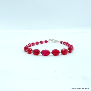 Puede incluir: Una pulsera con cuentas rojas translúcidas facetadas que se alternan con pequeños espaciadores metálicos de tono plateado. La pieza incluye un cierre de mosquetón de metal plateado y lleva el texto THE GEMSBEADS en la esquina.
