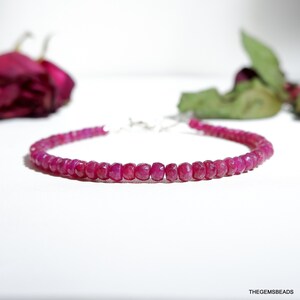 May include: A delicate bracelet featuring small, faceted, ruby gemstones strung together. The vibrant pink-red beads are connected with a silver clasp. A blurred background includes a dark red rose and green leaves.
