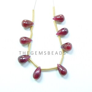 May include: A strand of eight teardrop-shaped gemstone beads strung on gold wire. The beads are a deep red color and have a glossy finish. The text "THE GEMSBeads" is visible in the image.