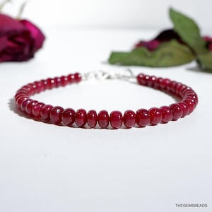 May include: A beaded bracelet featuring small, round, deep red beads. The beads are strung together with a silver-colored clasp. The bracelet is displayed on a white surface, with a blurred background that includes a rose.