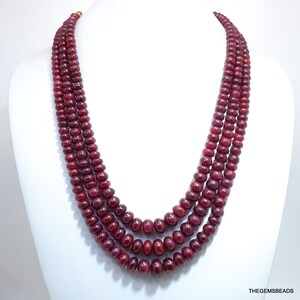 Puede incluir: Collar de varias hebras con filas de cuentas redondas de color rojo intenso. Las cuentas están ensartadas, creando un efecto de capas. El collar se exhibe en un maniquí blanco, mostrando el diseño y el color de la joya.