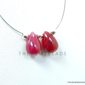 May include: Two teardrop-shaped ruby red gemstone beads with silver caps and loops. The beads are hanging on a silver wire. TheGEMSBeads is printed below the beads.