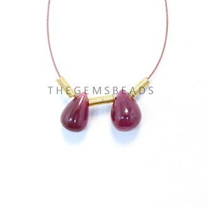 May include: Two teardrop-shaped ruby gemstones strung on a gold wire with gold spacers. The gemstones are a deep red color and are about 1 cm long. The wire is about 15 cm long. The text "THE GEMSBeads" is visible in the image.